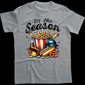 May include: Grey t-shirt with a baseball-themed design. The graphic features a baseball cap, bats, peanuts in a bucket, a hot dog, and a baseball. The text on the shirt reads "'Tis the Season".