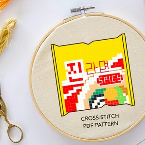 May include: A cross-stitch pattern for a yellow and red package of instant noodles with the text "Spicy" on it. The pattern is on a white fabric in a wooden embroidery hoop. The text "Cross-Stitch PDF Pattern" is below the image.