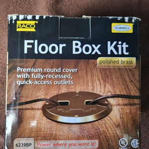 May include: A box for a floor box kit in polished brass. The box has a picture of the floor box kit installed in a wood floor. The text on the box reads "RACO Floor Box Kit polished brass Premium round cover with fully-recessed, quick-access outlets 6239BP Power where you want it!" and has UL listed and SP symbols.