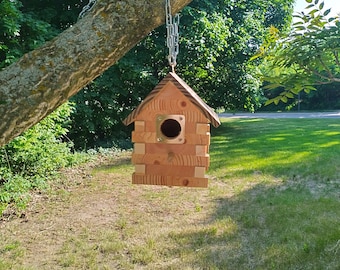 Può includere: Un nido di uccelli in legno con un foro di ingresso rotondo, appeso a un ramo di albero con una catena argentata. Il nido di uccelli è fatto di legno marrone chiaro e ha un design semplice e rustico.