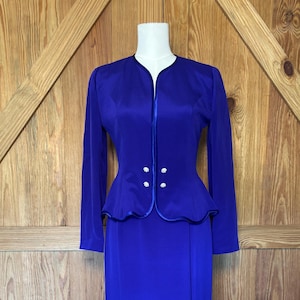 May include: A striking blue two-piece suit. The jacket features a peplum waist, long sleeves, and decorative buttons. The matching skirt is floor-length with a side slit. The suit is displayed on a mannequin.