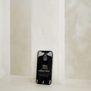 May include: A smartphone displaying a black and white image of a person wearing a blazer and crop top. The screen features the text client love in a cursive font, followed by smaller, less legible text below the main heading on the display.