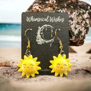 May include: A pair of gold dangle earrings with a yellow glitter sun charm. The sun has a smiling face with pink cheeks. The earrings are on a black card with the text "Whimsical Wishes".
