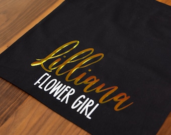Puede incluir: Bolsa de lona negra con letras doradas que dicen "Lilliana Flower Girl".