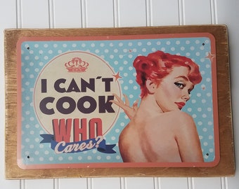 May include: A vintage-style metal sign with a blue and white polka dot background. The sign features a woman with red hair and the text "I can't cook who cares?" in pink and white lettering.