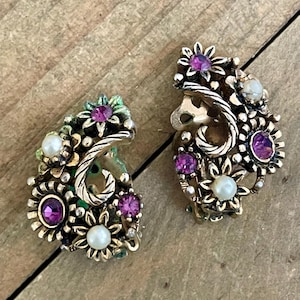 May include: A pair of vintage clip-on earrings. Each earring features a gold-toned metal floral design with purple and white gemstones. The earrings have a detailed, ornate appearance, with a mix of flower and swirl elements.