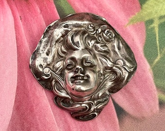May include: A silver brooch featuring a detailed sculpted face of a woman with flowing hair and a rose. The brooch is in the Art Nouveau style.