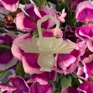 May include: A light green, carved glass basket-shaped ornament with a handle and a floral design, set against a backdrop of pink and white carnation flowers. The ornament has a gold ring attached to it.