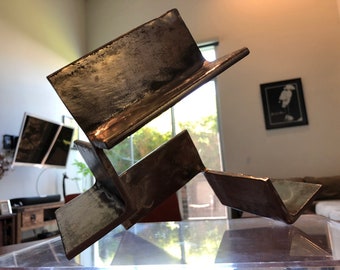 May include: A modern abstract sculpture made of metal. The sculpture is composed of four pieces of metal that are welded together. The metal is a dark brown color and has a rough, textured surface. The sculpture is sitting on a clear glass surface.