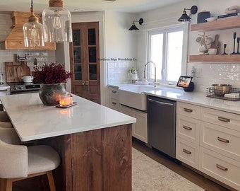 May include: A white farmhouse kitchen with a large island featuring a butcher block countertop and white quartz countertop. The kitchen has white cabinets with gold hardware, a farmhouse sink, and a stainless steel dishwasher. The kitchen also has a wood-paneled range hood and a white subway tile backsplash. The text "Northern Edge Woodshop Co." is visible on the backsplash.