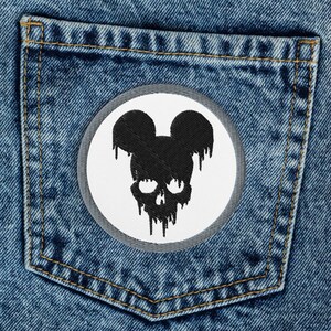 Pode incluir: Um par de calças de ganga azuis com um remendo branco no bolso de trás. O remendo apresenta uma silhueta preta de uma caveira com orelhas do Mickey Mouse.