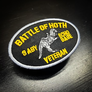 Puede incluir: Un parche ovalado negro con texto amarillo y un borde bordado blanco y gris. El texto dice "BATTLE OF HOTH", "ECHO BASE", "9 ABY" y "VETERAN". Una ilustraci&oacute;n blanca de una criatura est&aacute; en el centro.