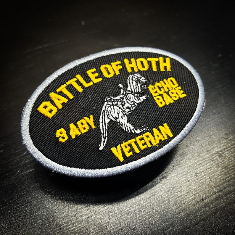 Puede incluir: Un parche ovalado negro con texto amarillo y un borde bordado blanco y gris. El texto dice "BATTLE OF HOTH", "ECHO BASE", "9 ABY" y "VETERAN". Una ilustraci&oacute;n blanca de una criatura est&aacute; en el centro.