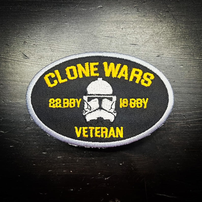 K&ouml;nnte beinhalten: Ein ovales Abzeichen mit den gelben Worten "CLONE WARS", &uuml;ber einer wei&szlig;en Helm-Grafik. Darunter die gelben Daten "22 BBY" und "19 BBY". Das Wort "VETERAN" steht unten in Gelb.