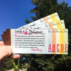 May include: A vintage Disneyland ticket book with a welcome message from Walt Disney. The ticket book is brown with a yellow, orange, and red striped border. The text reads "Welcome to Disneyland U.S.A."