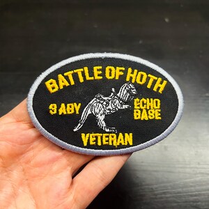 Puede incluir: Un parche ovalado con el texto amarillo "BATTLE OF HOTH", sobre una ilustraci&oacute;n en blanco y negro de una criatura. El parche tambi&eacute;n incluye el texto "9 ABY", "ECHO BASE" y "VETERAN" en amarillo. El parche tiene un fondo negro y un borde gris.