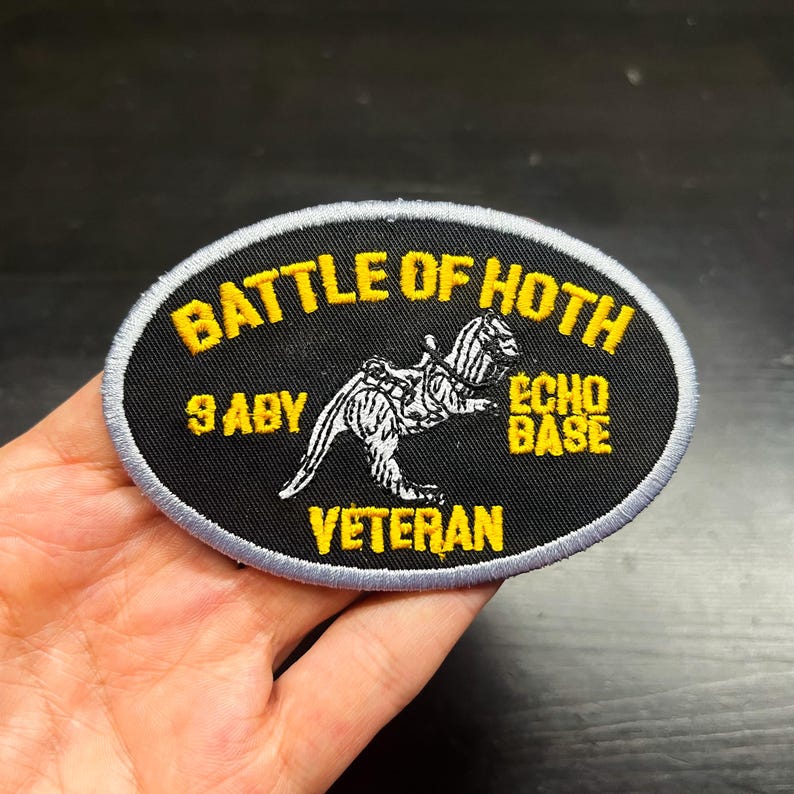 Puede incluir: Un parche ovalado con el texto amarillo "BATTLE OF HOTH", sobre una ilustraci&oacute;n en blanco y negro de una criatura. El parche tambi&eacute;n incluye el texto "9 ABY", "ECHO BASE" y "VETERAN" en amarillo. El parche tiene un fondo negro y un borde gris.