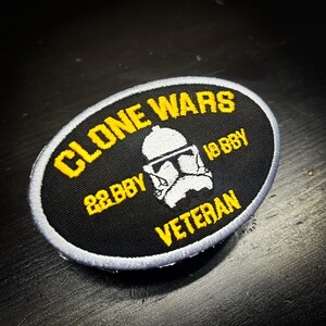 K&ouml;nnte beinhalten: Ovaler schwarzer Aufn&auml;her mit gelbem Text und einer wei&szlig;en Helm-Grafik. Der Text lautet "CLONE WARS", "22 BBY", "18 BBY" und "VETERAN". Der Aufn&auml;her hat einen grauen Rand.