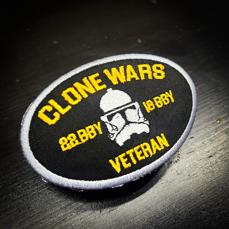 K&ouml;nnte beinhalten: Ovaler schwarzer Aufn&auml;her mit gelbem Text und einer wei&szlig;en Helm-Grafik. Der Text lautet "CLONE WARS", "22 BBY", "18 BBY" und "VETERAN". Der Aufn&auml;her hat einen grauen Rand.