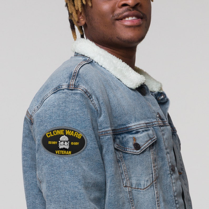 K&ouml;nnte beinhalten: Hellblaue Jeansjacke mit wei&szlig;em Kunstfellkragen. Die Jacke hat einen schwarzen Aufn&auml;her auf dem linken &Auml;rmel mit den Worten "CLONE WARS" und "VETERAN" sowie eine Grafik eines Sturmtrupplerhelms.