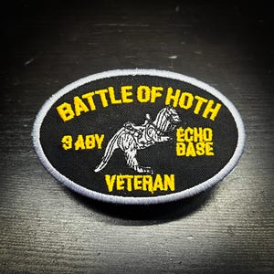 May include: An oval black patch with yellow text. The text reads "BATTLE OF HOTH", "ECHO BASE", "9 ABY", and "VETERAN". A white and black illustration of a creature is in the center of the patch.