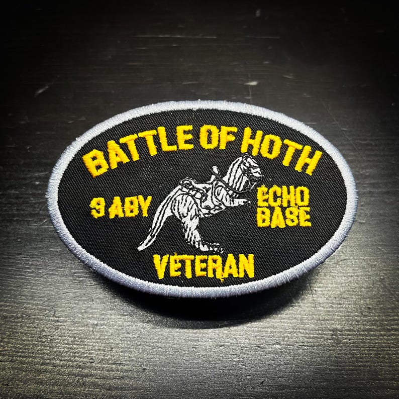 Puede incluir: Un parche ovalado negro con texto amarillo. El texto dice "BATTLE OF HOTH", "ECHO BASE", "9 ABY" y "VETERAN". En el centro hay una ilustraci&oacute;n en blanco y negro de una criatura.