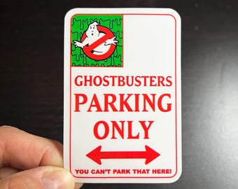 May include: A white sign with red text that says "Ghostbusters Parking Only" with a red arrow pointing left and right. The sign also has a green and white image of a ghost with a red circle and line through it. The text at the bottom of the sign says "You can't park that here!"