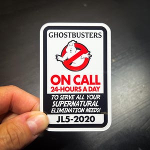 May include: A white sticker with black and red text that reads "GHOSTBUSTERS ON CALL 24-HOURS A DAY TO SERVE ALL YOUR SUPERNATURAL ELIMINATION NEEDS! JL5-2020". The sticker features a cartoon ghost with a red circle and slash through it.