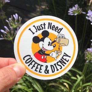 May include: A round sticker with a yellow border and a white background. The sticker features a cartoon image of Mickey Mouse pouring coffee from a pot into a cup. The text "I Just Need Coffee & Disney" is written in a black circle around Mickey Mouse.