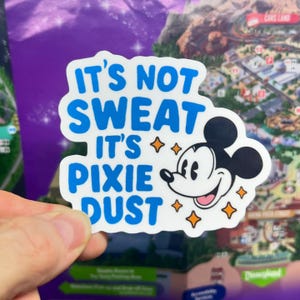 May include: A white sticker with blue text that reads "IT'S NOT SWEAT IT'S PIXIE DUST." The sticker features a smiling Mickey Mouse and orange star accents. The sticker is held in front of a map.