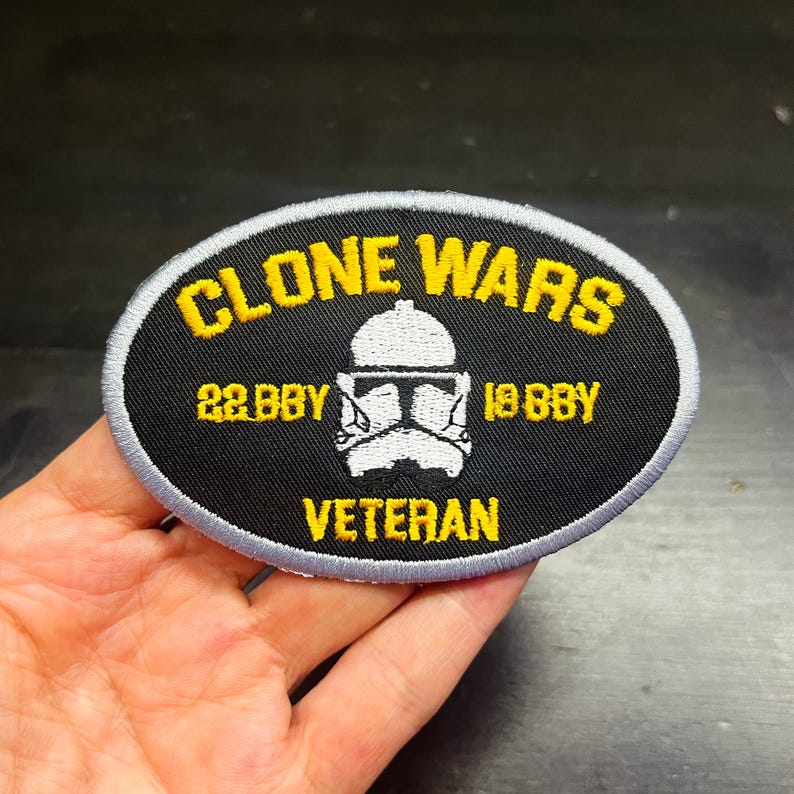 K&ouml;nnte beinhalten: Ein ovales Abzeichen mit den gelben Worten "CLONE WARS", einer wei&szlig;en Helm-Grafik und dem Wort "VETERAN" in Gelb. Das Abzeichen hat einen schwarzen Hintergrund mit einem grauen Rand.