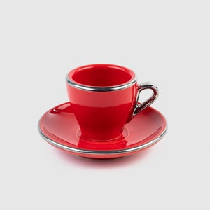 Könnte beinhalten: Ein leuchtend rotes Espresso-Tassen- und Untertassen-Set. Die Tasse und die Untertasse haben eine glänzende Oberfläche und sind mit einem silberfarbenen Metallband versehen. Die Tasse hat einen passenden silbernen Griff. Das Set ist für Espresso oder Kaffee gedacht.