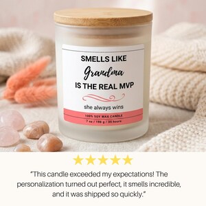 K&ouml;nnte beinhalten: Duftkerze in einem mattierten Glas mit Holzdeckel. Das Etikett lautet "SMELLS LIKE Grandma IS THE REAL MVP" mit "she always wins" darunter. Die Kerze hat 198 g und brennt 35 Stunden. Rosa Akzente und Steine sind sichtbar.