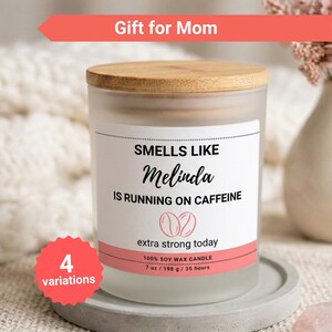 K&ouml;nnte beinhalten: Eine Kerze aus mattiertem Glas mit einem Holzdeckel. Auf dem Etikett steht "Smells Like Melinda Is Running on Caffeine". Die Kerze ist 198 g und besteht aus 100 % Sojawachs. Der Text "Gift for Mom" steht dar&uuml;ber.