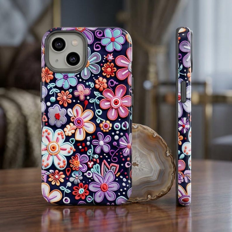 Op de afbeelding: Een donkerblauwe telefoonhoes met een verhoogd bloemenontwerp in tinten roze, paars, oranje en blauw. De hoes heeft een matte afwerking en een beschermende bumper rond de camera. De zijkant van de hoes heeft ook het bloemenpatroon.