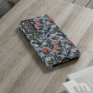 Op de afbeelding: Een blauwe telefoonhoes met een bloemenpatroon. De bloemen zijn roze, blauw en geel en steken uit de oppervlakte van de hoes.