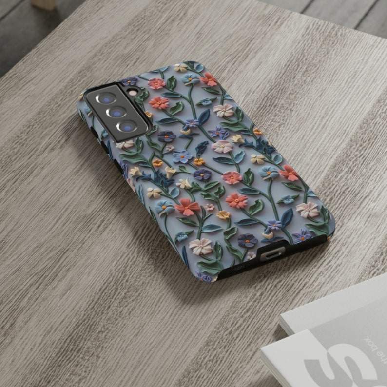 Op de afbeelding: Een blauwe telefoonhoes met een bloemenpatroon. De bloemen zijn roze, blauw en geel en steken uit de oppervlakte van de hoes.