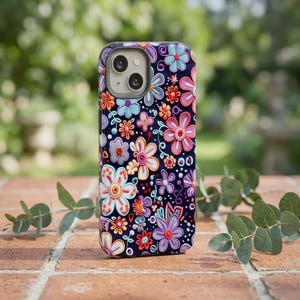 Op de afbeelding: Een telefoonhoesje met een levendig bloemenpatroon. De hoes heeft een donkerblauwe achtergrond, versierd met kleurrijke bloemen in roze, oranje, paars en blauw. De bloemen hebben gedetailleerde bloemblaadjes en zijn gerangschikt in een herhalend patroon.
