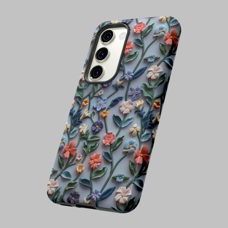 Op de afbeelding: Een blauwe telefoonhoes met een bloemenontwerp met roze, blauwe en witte bloemen. De hoes heeft een verhoogde camerabult met drie lenzen.
