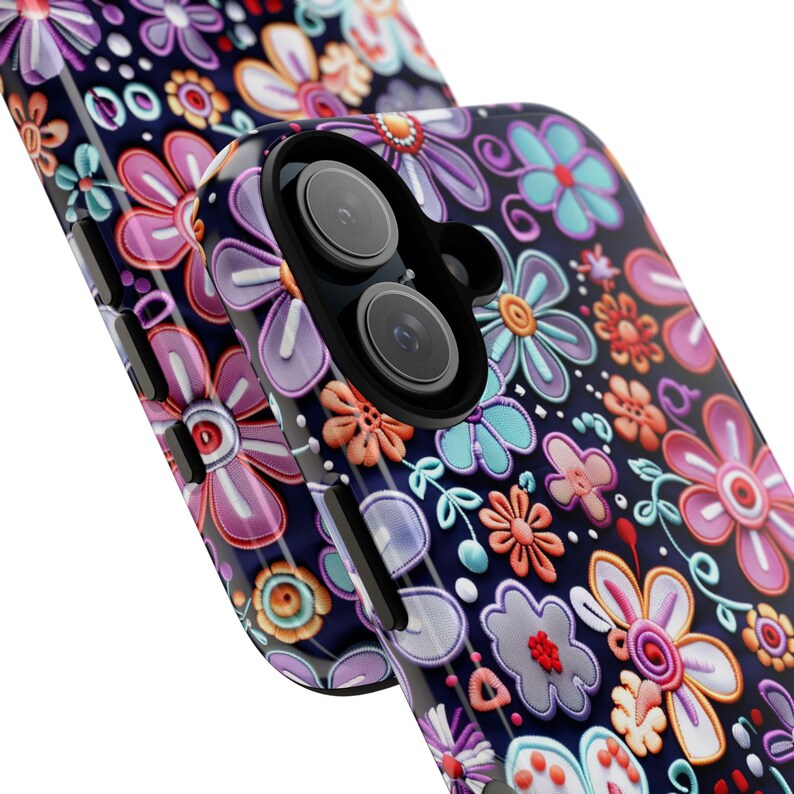 Op de afbeelding: Close-up van twee telefoonhoesjes met een bloemenpatroon. De hoesjes zijn donkerblauw met een verhoogd, kleurrijk patroon van bloemen in roze, paarse, oranje en blauwgroene tinten. De telefoonhoes aan de rechterkant heeft twee cameralenzen.