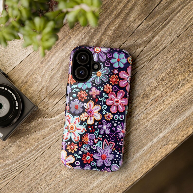 Op de afbeelding: Een smartphonehoesje met een bloemenpatroon. De hoes heeft een donkere achtergrond met een herhalend ontwerp van kleurrijke bloemen in roze, paars, blauw en oranje. De hoes heeft een glanzende afwerking en is ontworpen voor een smartphone.