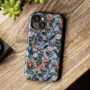 Op de afbeelding: Een telefoonhoesje met een bloemenprint. Het hoesje is blauw met witte bloemen en groene bladeren. De cameralenzen zijn zichtbaar op de achterkant van het hoesje.