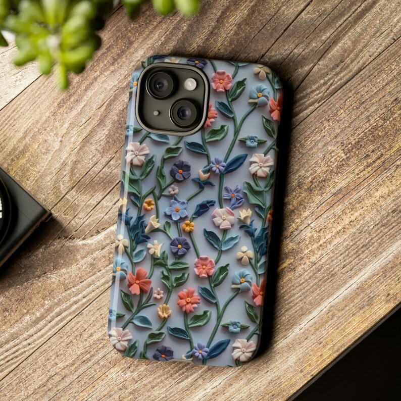 Op de afbeelding: Een telefoonhoesje met een bloemenprint. Het hoesje is blauw met witte bloemen en groene bladeren. De cameralenzen zijn zichtbaar op de achterkant van het hoesje.