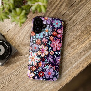 Op de afbeelding: Een smartphonehoesje met een kleurrijk bloemenpatroon. De hoes heeft een donkere achtergrond met diverse bloemen in roze, paars, blauw, oranje en wit. Het ligt op een houten oppervlak.