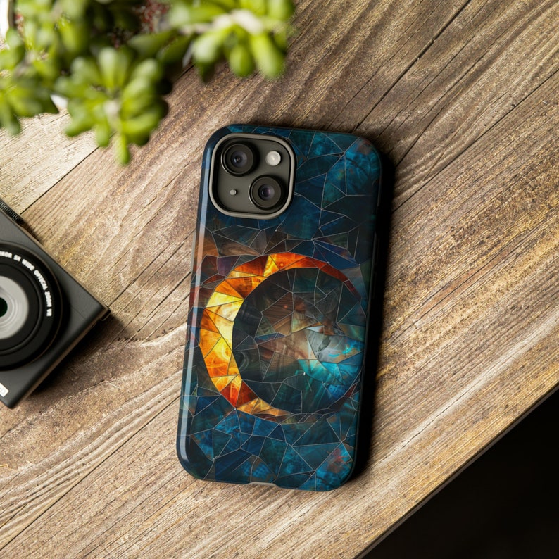 May include: A phone case with a mosaic design featuring a crescent moon with a sun-like pattern inside. The case is blue, green, and orange.