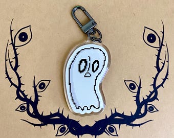 May include: A clear acrylic keychain featuring a white pixel art ghost with a surprised expression, attached to a dark metal lobster clasp. The ghost has a black outline and simple facial features, including two oval eyes and a small mouth.