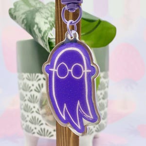 Puede incluir: Un llavero morado con forma de fantasma con contornos blancos y gafas. El llavero está sujeto a un cierre morado. El fondo incluye una planta en una maceta decorativa y un soporte de madera.