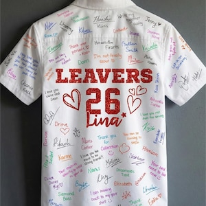 Peut inclure: Chemise blanche &agrave; manches courtes orn&eacute;e de signatures manuscrites multicolores. Le texte "LEAVERS 26 Lina" en paillettes rouges est &eacute;galement pr&eacute;sent. Des c&oelig;urs et des &eacute;toiles sont dessin&eacute;s, probablement un article comm&eacute;moratif.