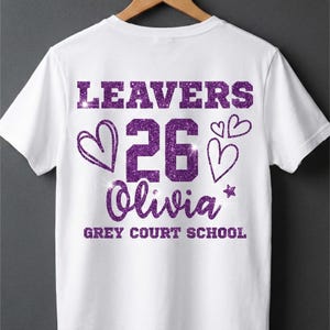 Peut inclure: T-shirt blanc avec texte paillet&eacute; violet &laquo;&nbsp;LEAVERS 26 Olivia&nbsp;&raquo; avec des graphiques de c&oelig;ur et d'&eacute;toile. En dessous du nom se trouve &laquo;&nbsp;GREY COURT SCHOOL&nbsp;&raquo;. Le t-shirt est suspendu &agrave; un cintre en bois.