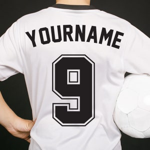 Può includere: Maglia da calcio bianca con testo nero che dice "YOURNAME" e il numero "9" sul retro.